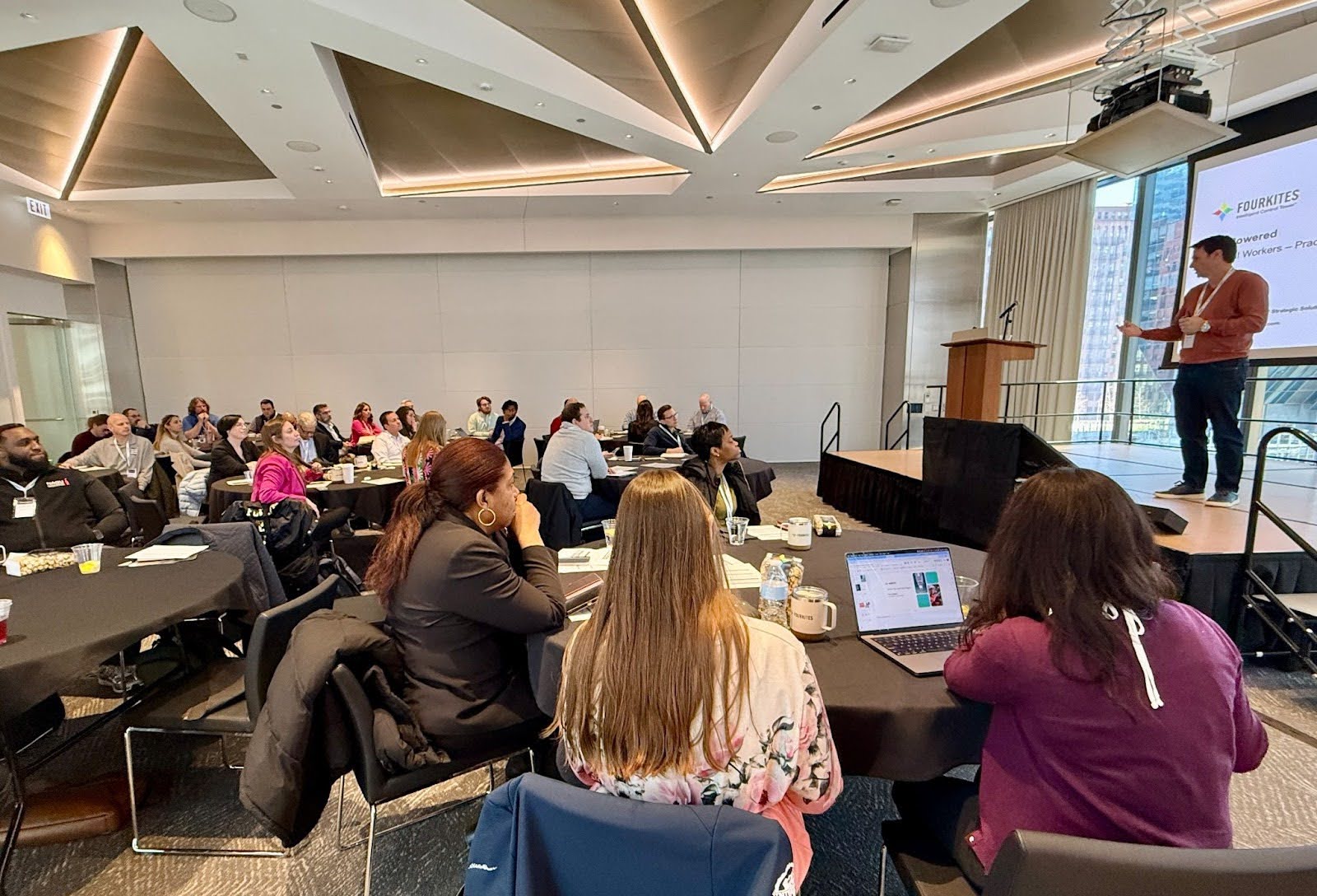 Conference room with speaker on stage presenting slides on a screen. Audience are sitting at roundtables with their laptops open.
