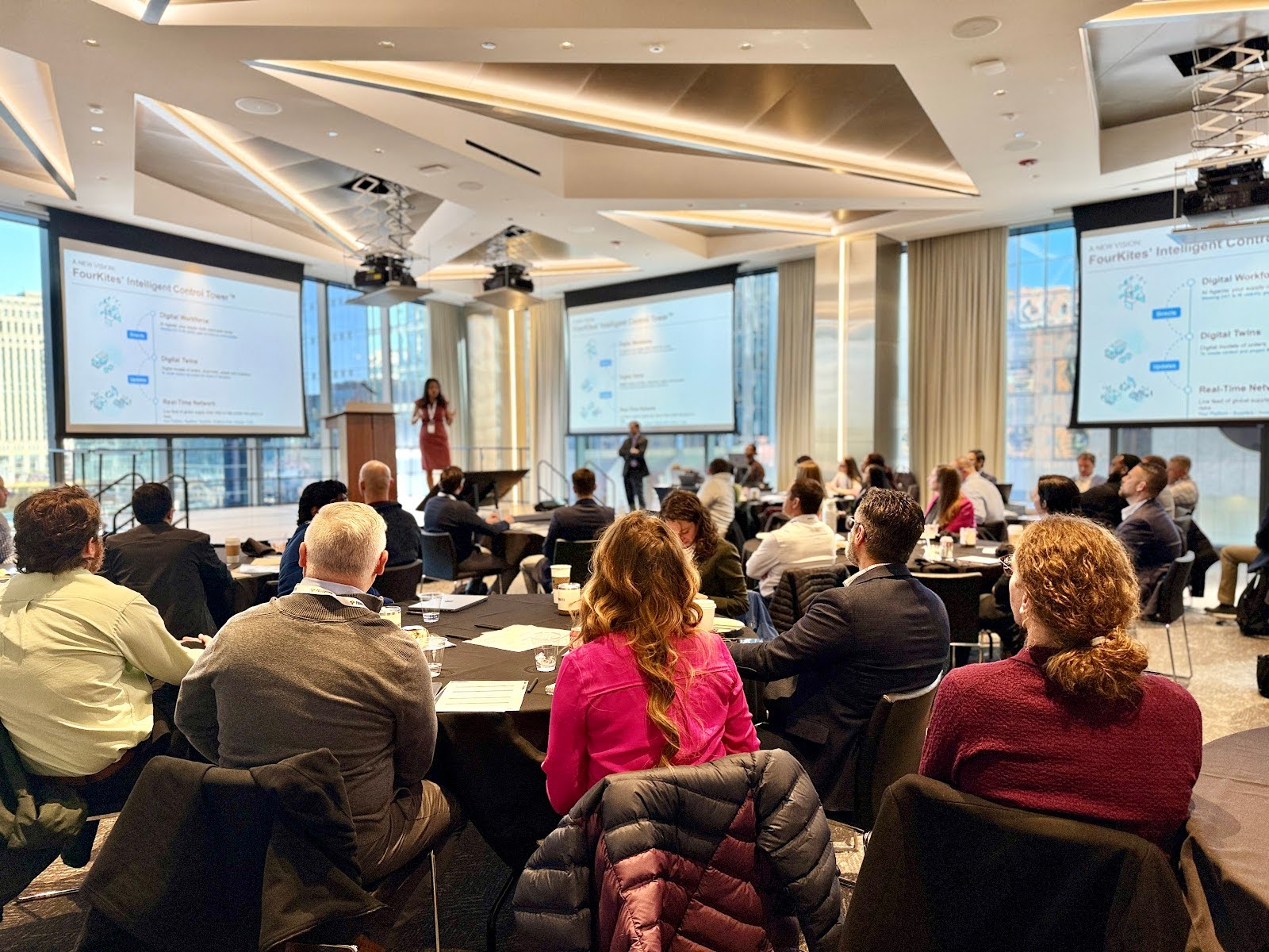 Conference room with speaker on stage presenting. Three screens surround the room and people sitting at roundtables as the audience.